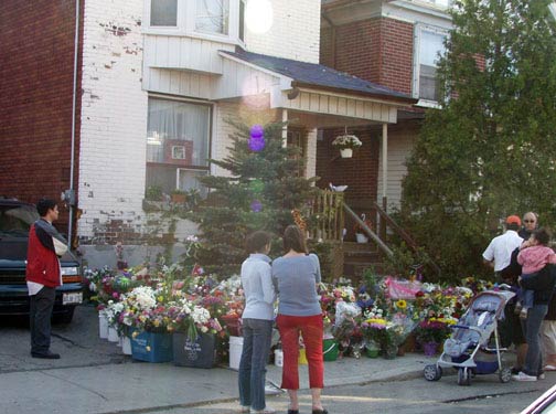 people gathered outside numerous bunches of flowers, in front of a house